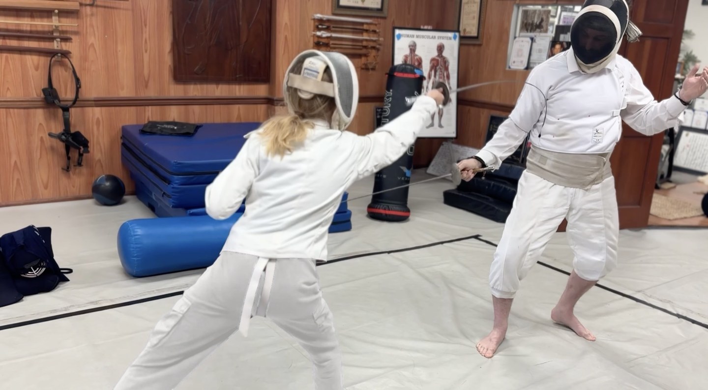 Two fencers in whites and masks facing off with epees in the training hall