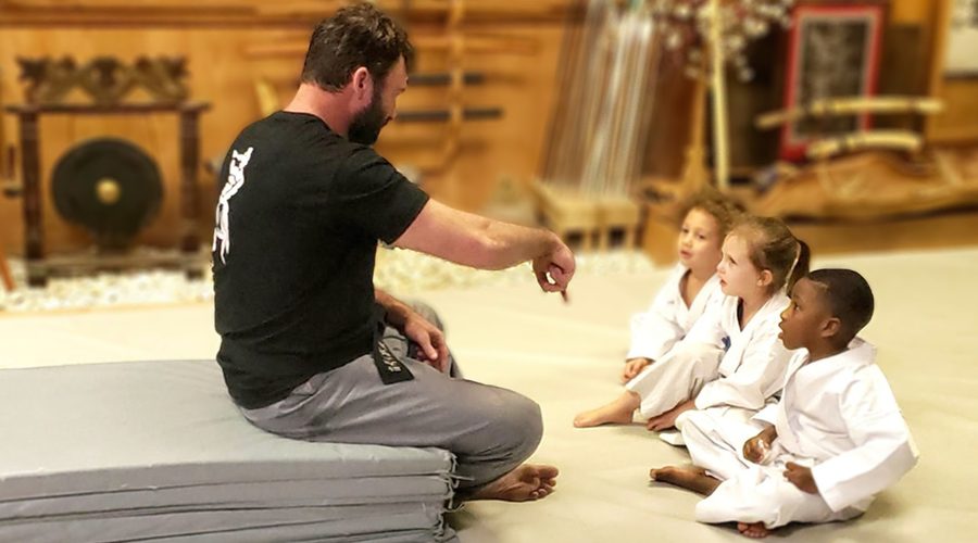 An instructor seated on the mat talking with three young students at Excalibur Academy