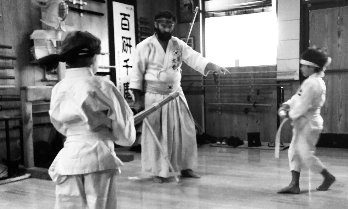 An instructor in hakama guiding two children in gi through a sword form